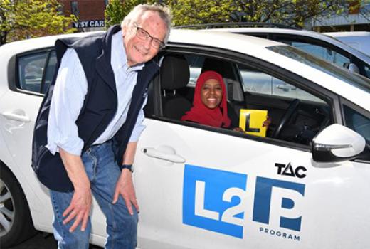 Young woman wearing hijab sitting inside car holding an L plate.  Older man next to car.  Car has L2P TAC program on the car door.