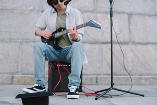 Young man sitting on an amplifier playing the guitar on a footpath with a hat upside down on the ground in front of him