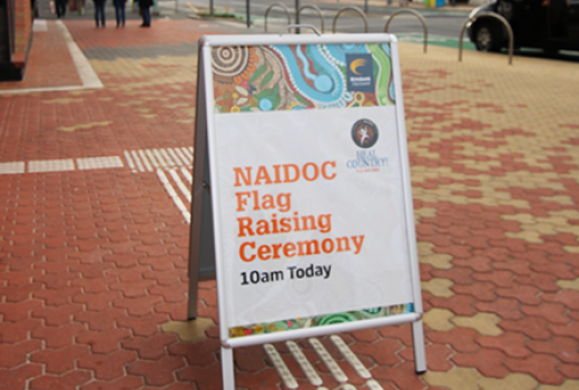 A frame sign on bricked pavement indicating NAIDOC Flag Raising Ceremony 10am Today