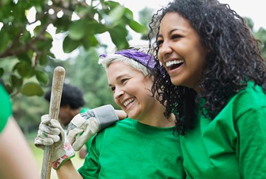 Two women wearing gardening gloves and hold garden tool and smiling together.