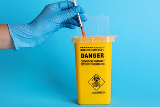 Gloved hand putting used syringe/ injection into a sharps container fmarked Danger