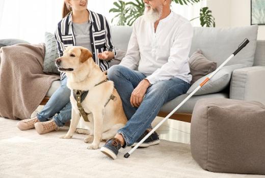 Older man sitting on couch next to young woman.  Guide dog sitting on the floor. White cane leaning against couch. 